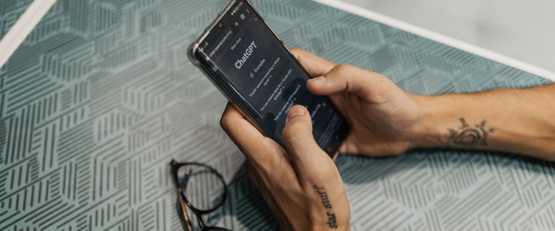 A person holding a phone showing the ChatGPT app open, glasses resting on the table beside them.